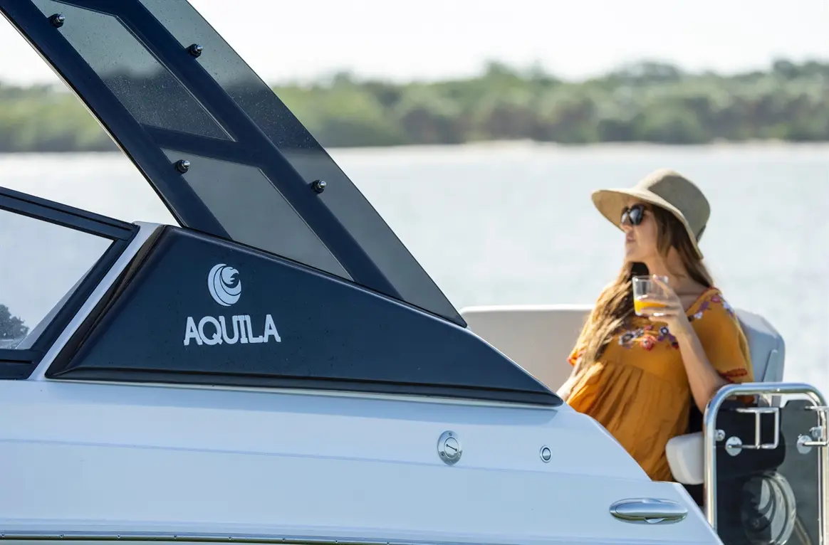 A woman in sunglasses and a sun hat enjoys a drink on an Aquila boat near the water, with greenery in the background.